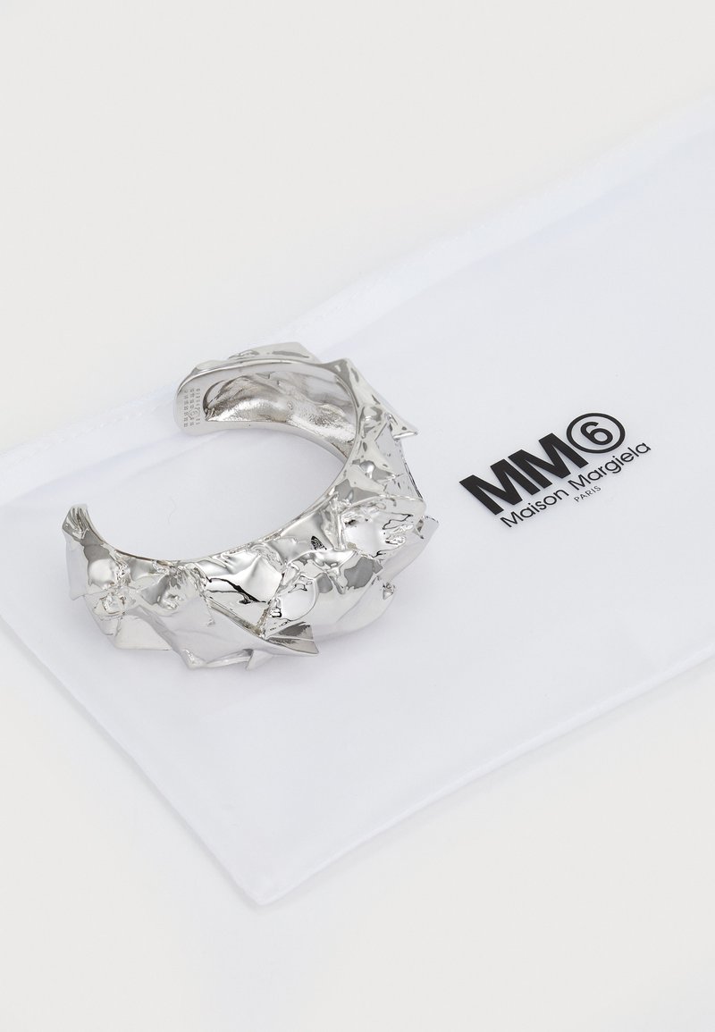Zilveren metalen armband met een sculpturale, scherpere vormgeving en een reflecterende afwerking, geplaatst op een zachte witte pouch met logo.
