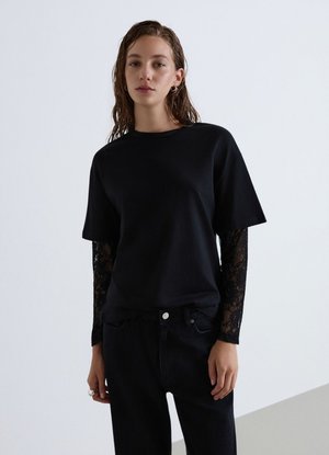Donna con capelli bagnati che indossa una maglietta nera a maniche corte sopra una camicia nera di pizzo a maniche lunghe e pantaloni neri su uno sfondo chiaro semplice.