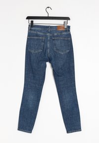 Tmavě modré džíny z denimu s úzkým střihem, zobrazující dvě zadní kapsy a koženou nášivku na pásku, zavěšené na černém ramínku.