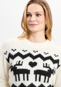 Strickpullover mit einem cremigen Grundton, verziert mit schwarzen Rentier- und Herzmustern, einem Zickzackdesign und einem runden Ausschnitt. Weiche Textur.