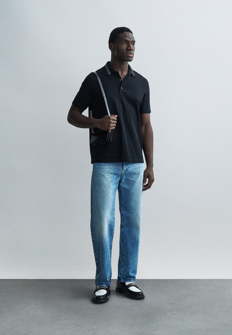 Homme portant un polo noir, un jean bleu avec des boutons sur le côté, des mocassins noir et blanc, tenant un sac à bandoulière noir, debout devant un mur uni.