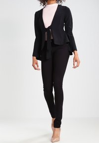Chaqueta peplum negra con un lazo en la parte delantera, llevada sobre una blusa rosa clara; combinada con pantalones ajustados negros y zapatos de tacón nude.