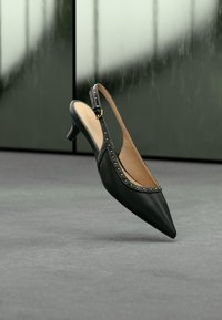 Chaussure à talon haut à bride arrière en cuir noir avec bout pointu et détails cloutés dorés, flottant au-dessus d'un sol gris.