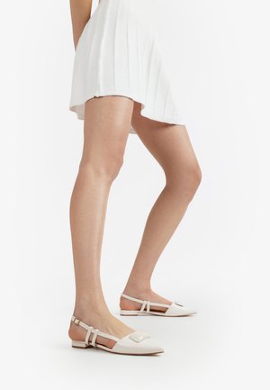 Frau trägt weiße spitze Slingback-Ballerinas mit doppelten Riemchen und einem weißen plissierten Minirock vor weißem Hintergrund.