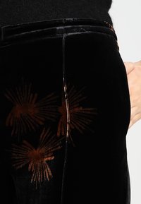 Zwarte fluwelen broek met bruine bloemenpatronen, een slank ontwerp en een gladde textuur, benadrukt door een subtiele glans langs de naden.