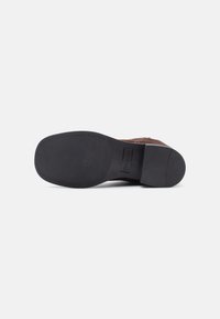 Chaussure en cuir marron avec un bout arrondi, surface texturée et une semelle en caoutchouc noir. Présente un branding subtil sur la semelle.