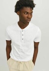 Fehér, rövid ujjú henley póló, három szürke gombbal, könnyű anyagból készült, bézs csíkos rövidnadrággal viselve. A modell kezét a zsebében tartva pózol.