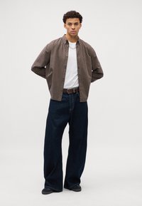 Camisa de botones a rayas marrones y blancas, camiseta blanca debajo, combinada con pantalones de mezclilla oscuros holgados y zapatos negros, de pie contra un fondo claro.