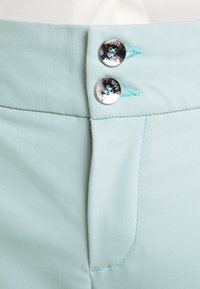 Ceinture de vêtement bleu clair avec deux boutons argentés brillants et un fil turquoise fixé sur un fond de tissu blanc.