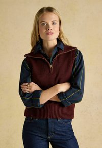 Maroon gestrickte Weste mit einem Reißverschlusskragen über einem dunkelgrünen Hemd mit horizontalen Streifen. Das Modell hat die Arme verschränkt und trägt einen lässigen Look.