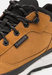 Tan suede sneaker with black mesh tongue, contrasting stitching, black laces, and a white and black rubber sole. Timberland logo tag visible.