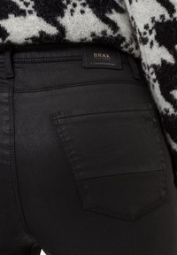 Jeans de mezclilla negra con un corte ajustado, que presentan un único bolsillo trasero y una etiqueta de BRAX, colocados sobre un suéter de patrón texturizado.