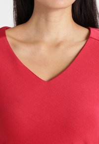 Top rosso con scollatura a V realizzato in tessuto liscio ed elasticizzato. Presenta un design semplice con un colletto pulito e cuciture minime.