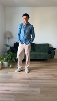 Denim srajca s gumbi, svetlo roza hlače in bele superge. Zelen kavč v ozadju z črnimi blazinami in lončnico na tleh.