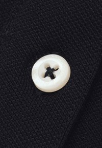 Czarna tkanina o fakturze z dużym białym guzikiem z czterema otworami oraz czarnymi detalami haftu. Guzik ma wytłoczone oznaczenie marki wokół krawędzi.