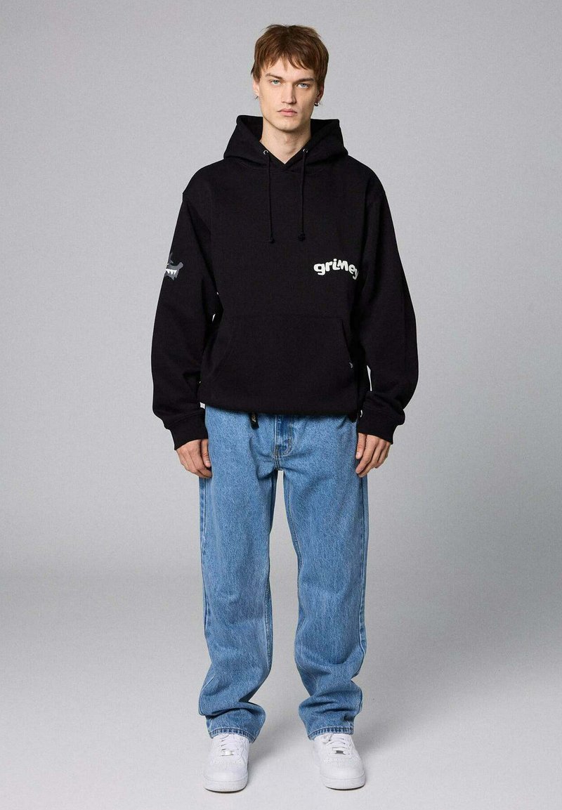 Felpa nera con tasca frontale e testo bianco, abbinata a jeans azzurri chiari e sneaker bianche. Il design presenta un logo sulla manica.