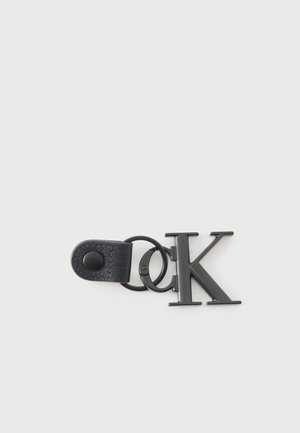 Fekete fém kulcstartó, amelyen összefonódott "c" és "k" betűk láthatók, egy kis fekete bőr pánttal, gombos záródással ellátva.