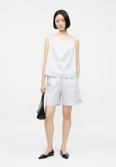 Γυναίκα με ανοιχτό μπλε ριγέ τοπ με λεπτές τιράντες και ταίριασμα σορτς, κρατώντας μια μαύρη τσάντα χειρός, φορώντας μαύρα μυτερά παπούτσια, στέκεται σε λευκό φόντο.