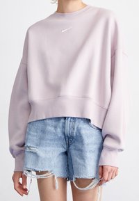 Osoba ubrana w jasnofioletową bluzę Nike i podarte jasnoniebieskie szorty jeansowe na jednolitym białym tle.