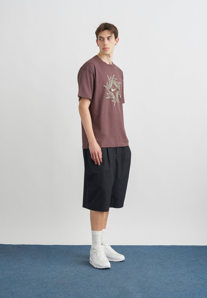 Junger Mann steht drinnen und trägt ein braunes T-Shirt mit Grafik, schwarze knielange Shorts, weiße Socken und weiße Turnschuhe auf blauem Teppich.