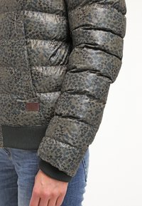 Kurtka pikowana w kamuflażowy wzór ciemnej zieleni i czerni, z teksturowaną powierzchnią, zapięciami na guziki oraz ściągaczami na mankietach.