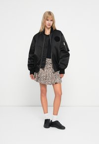 Blouson aviateur noir avec un patch rond, associé à une jupe à imprimé léopard. Le mannequin porte des baskets noires et des chaussettes blanches.