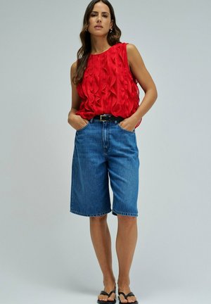 Mujer con blusa roja sin mangas con volantes, pantalones cortos de mezclilla azules hasta la rodilla de cintura alta, cinturón negro y sandalias negras, de pie con las manos en los bolsillos.