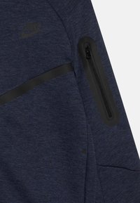 Námornícky modrá Nike bunda s textúrovanou látkou, má čiernu zipsovú kapsu na rukáve a čierny horizontálny pás cez hrudník.