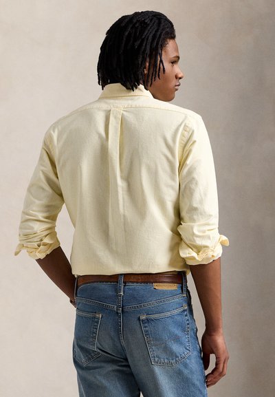 Κίτρινο ριγέ πουκάμισο με κουμπιά και μανίκια γυρισμένα, συνδυασμένο με μπλε τζιν τζιν. Σημαντικές λεπτομέρειες περιλαμβάνουν μια καφέ δερμάτινη ζώνη και τσέπες.