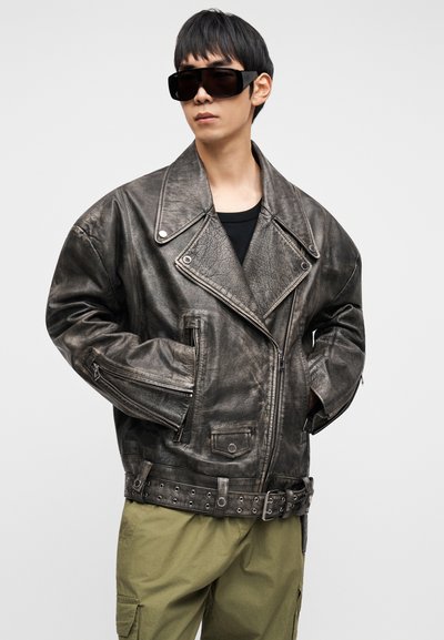 Γκρι δερμάτινο μπουφάν μοτοσικλέτας με μεγάλους γιακάδες, τσέπες με φερμουάρ και ζώνη στη μέση. Φορεμένο πάνω από μαύρο τοπ και συνδυασμένο με χακί παντελόνι.
