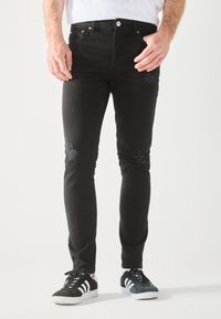 Muž v čiernych roztrhnutých skinny nohaviciach a čierno-bielych teniskách, stojaci proti jednoduchému svetlému pozadiu.