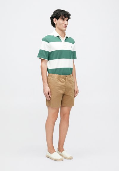 Polo cu dungi verzi și albe, pantaloni scurți kaki și espadrile de culoare deschisă. Are mâneci scurte, guler deschis și talie elastică.