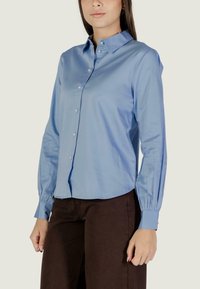 Donna che indossa una camicia blu chiaro con bottoni di perla e maniche lunghe abbinata a pantaloni marrone scuro, in piedi davanti a uno sfondo semplice.