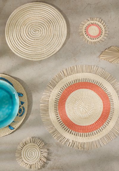 Set of woven round placemats in natural fibers. Features include soft beige, red accents, fringe details, and a large turquoise bowl.