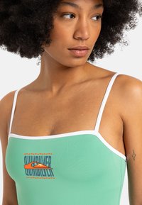 Grön bikiniöverdel med vita remmar, prydd med en framträdande "Quiksilver"-logotyp i orange mot en texturerad bakgrund. Enkel och strömlinjeformad design.