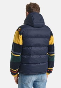 Navy Puffer-Weste mit Kapuze, ausgestattet mit mehrfarbigen Fleecemanschetten in gelben, grünen, blauen und roten Streifen, strukturierter und gepolsterter Design.