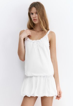 Jeune femme aux longs cheveux portant une robe blanche sans manches avec un col à volants et une jupe froncée, debout sur fond blanc.