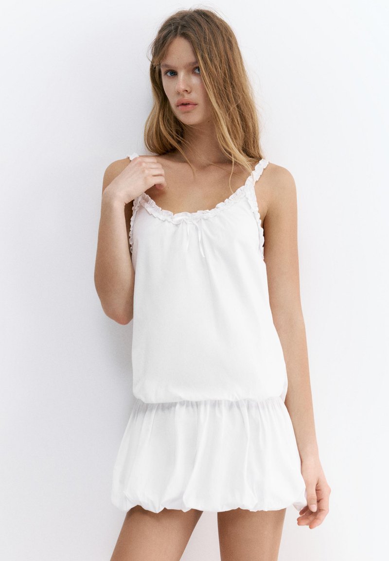 Jeune femme aux longs cheveux portant une robe blanche sans manches avec un col à volants et une jupe froncée, debout sur fond blanc.