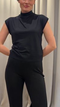 Woman standing with hands behind back wearing a black sleeveless turtleneck top and black pants against a neutral curtain backdrop.