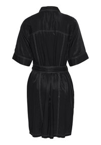 Melnas satīna kleita ar īsām piedurknēm, pogām vērstā apkakle un josta viduklī. Iezīmējas ar baltu kontrastējošu šuvumu un plūstošu apakšējo malu.