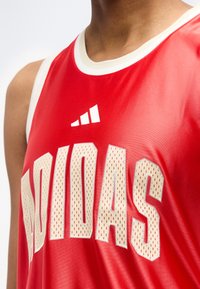 Κοντινό πλάνο ενός ατόμου που φοράει κόκκινη αθλητική φανέλα Adidas με λευκή επένδυση και μεγάλα μπεζ διάτρητα γράμματα.