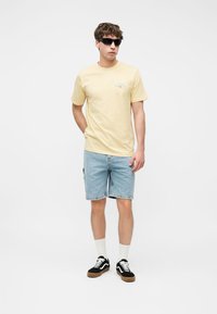 Uomo con occhiali da sole neri, maglietta giallo chiaro, pantaloncini di denim, calzini bianchi e scarpe da ginnastica nere in piedi contro uno sfondo semplice.