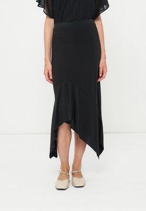 Femme portant une jupe midi asymétrique noire avec des chaussures plates beiges attachées aux chevilles, debout sur un fond blanc uni.