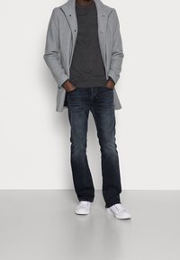 Harmaa villatakki yllään tumma neule, yhdistetty sinisiin farkkuihin ja valkoisiin tennareihin. Takissa on jämäkkä kaulus ja napit yksityiskohdissa.