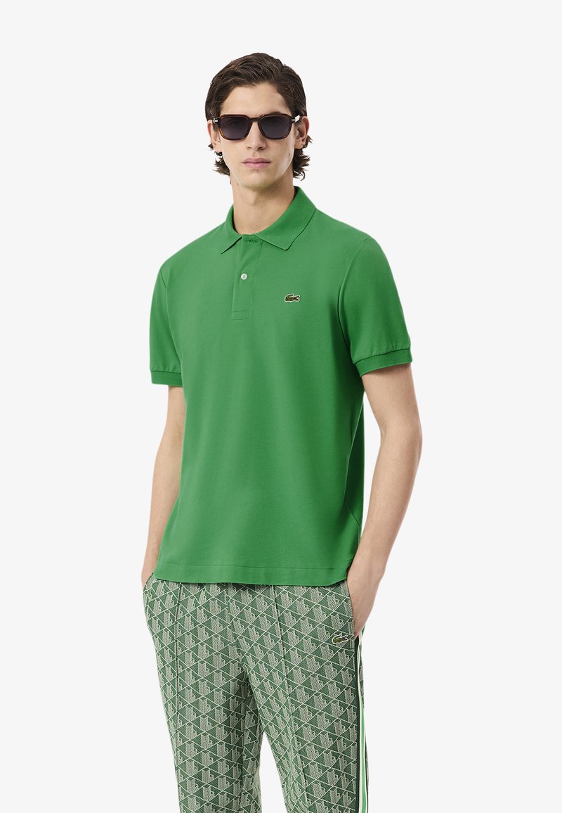 Polo verde con colletto classico, maniche corte e un piccolo logo, abbinato a pantaloni verdi con motivo e accenti bianchi.