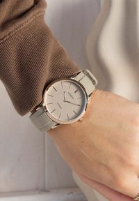 Montre ronde en argent avec un cadran beige, des marquages minimalistes et un bracelet en silicone gris clair. Aiguille positionnée sur une manche en cuir brun texturé.