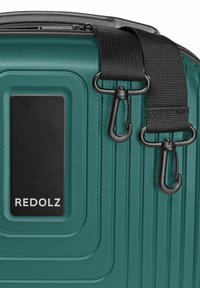 Teal harde kofferhoek met een gestructureerde oppervlakte, zwart stoffen riem met twee zwarte metalen draaikoppelingen, en een zwart label met de tekst "REDOLZ."
