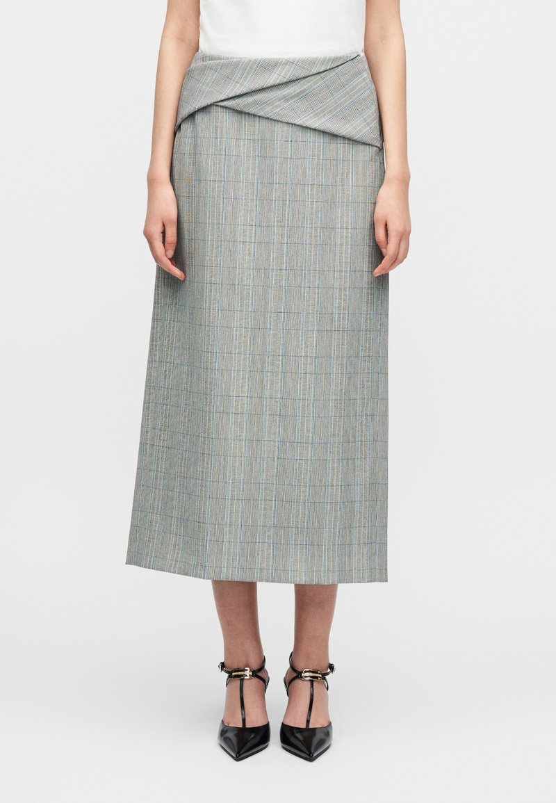 Γυναίκα που φοράει midi φούστα σε ψηλή μέση με γκρι καρό σχέδιο, ζώνη διπλωμένη στη μέση και μαύρες γόβες με μυτερή μύτη και λουράκι σε σχήμα Τ.