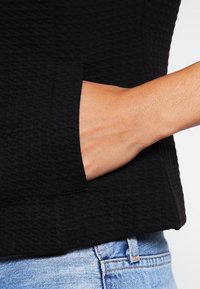 Haut noir texturé avec une main visible dans la poche. Le matériau semble tissé ; présente un ourlet droit et une coupe décontractée, associé à un denim bleu clair.