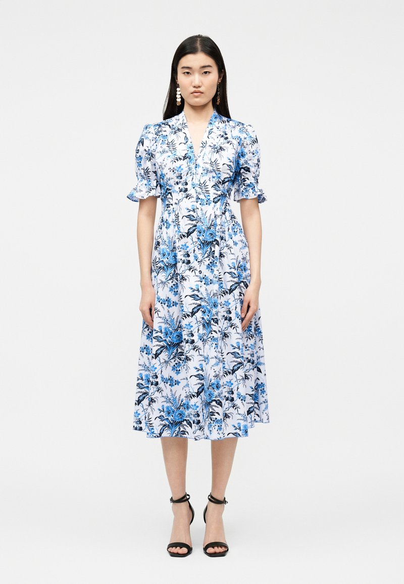 Γυναίκα που φοράει λευκό μεσοφόρεμα με μπλε λουλουδάτο σχέδιο, μανίκια φουσκωτά και μαύρα πέδιλα με λουράκι στον αστράγαλο, στέκεται μπροστά σε απλό ανοιχτό φόντο.